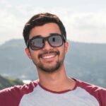 Young man in sunglasses leaning on a balcony with a scenic city view in the background.